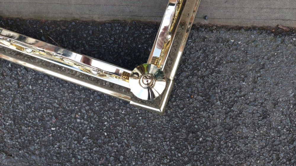 A polished brass extending fireplace fender featuring decorative side gallery rails with scrollwork and urn-shaped corner finials. View V