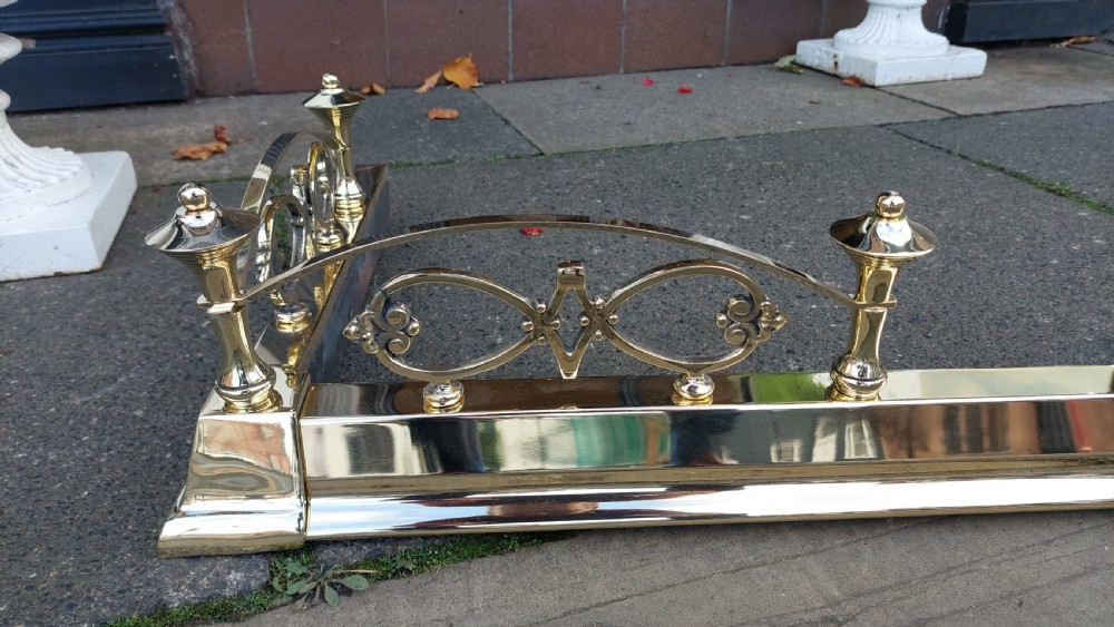 A polished brass extending fireplace fender featuring decorative side gallery rails with scrollwork and urn-shaped corner finials. View VI