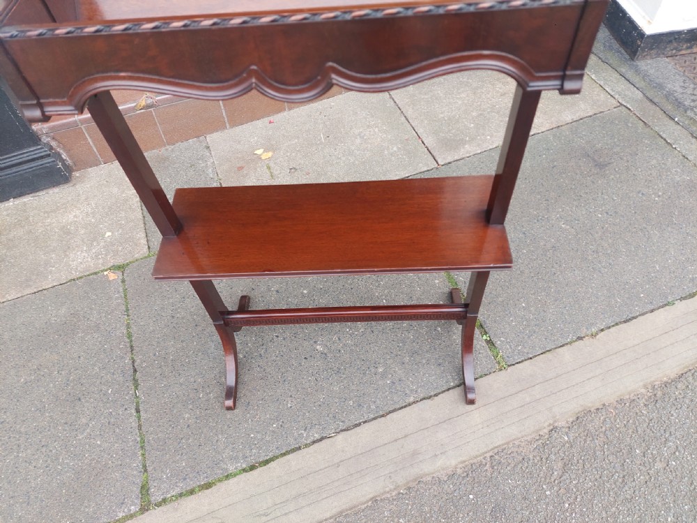 An Edwardian mahogany two-tier standing book trough featuring a shaped top apron, middle shelf, carved stretcher, and splayed legs. View IV