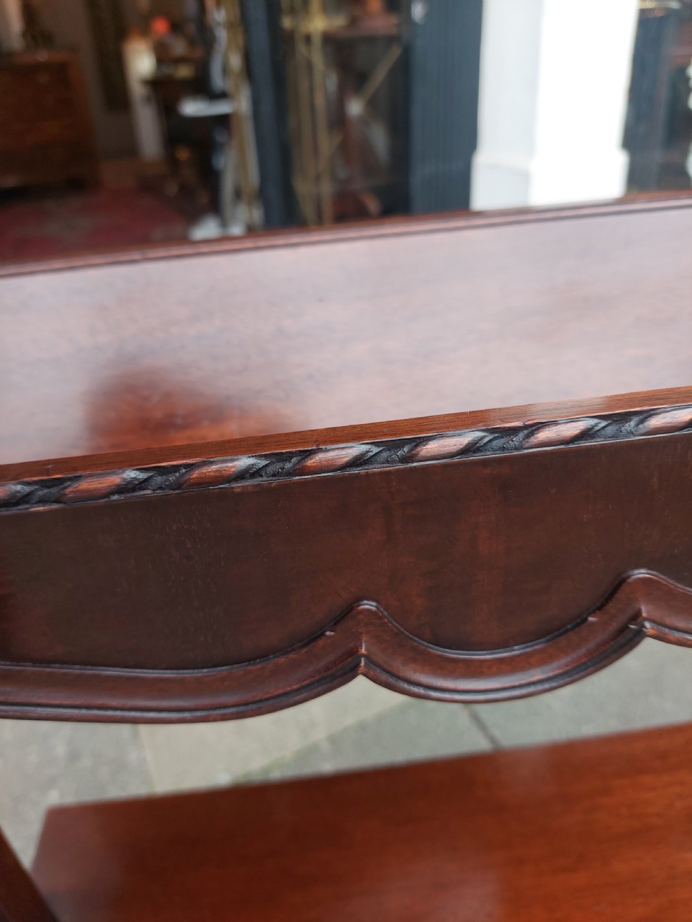 An Edwardian mahogany two-tier standing book trough featuring a shaped top apron, middle shelf, carved stretcher, and splayed legs. View VII