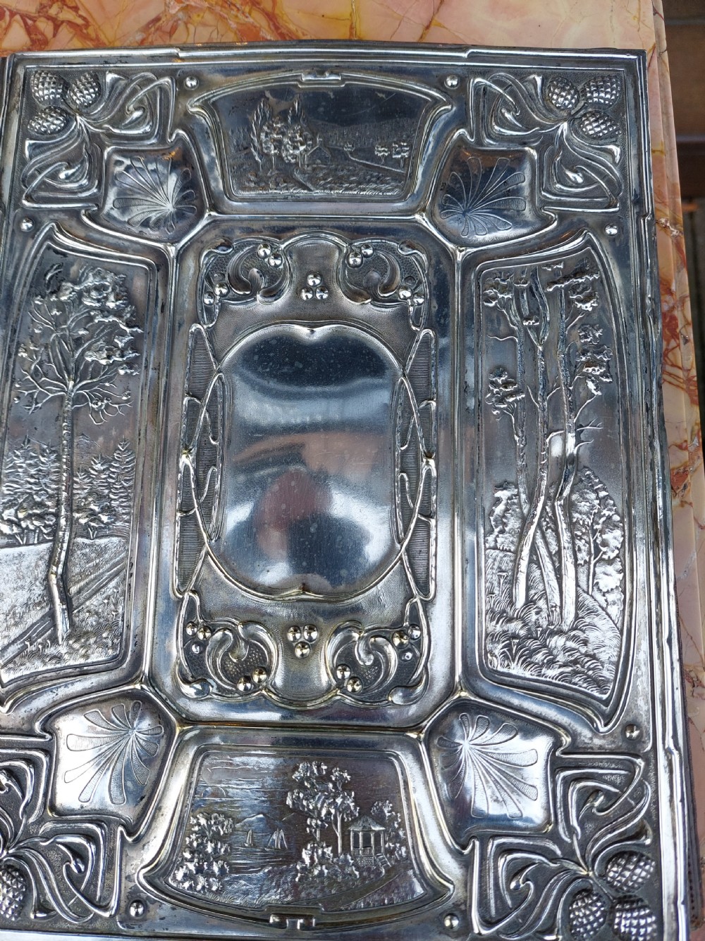 A rectangular silver-plated Art Nouveau blotter folio featuring relief embossed panels of trees, landscapes, pinecone motifs, and a central polished cartouche. View VII
