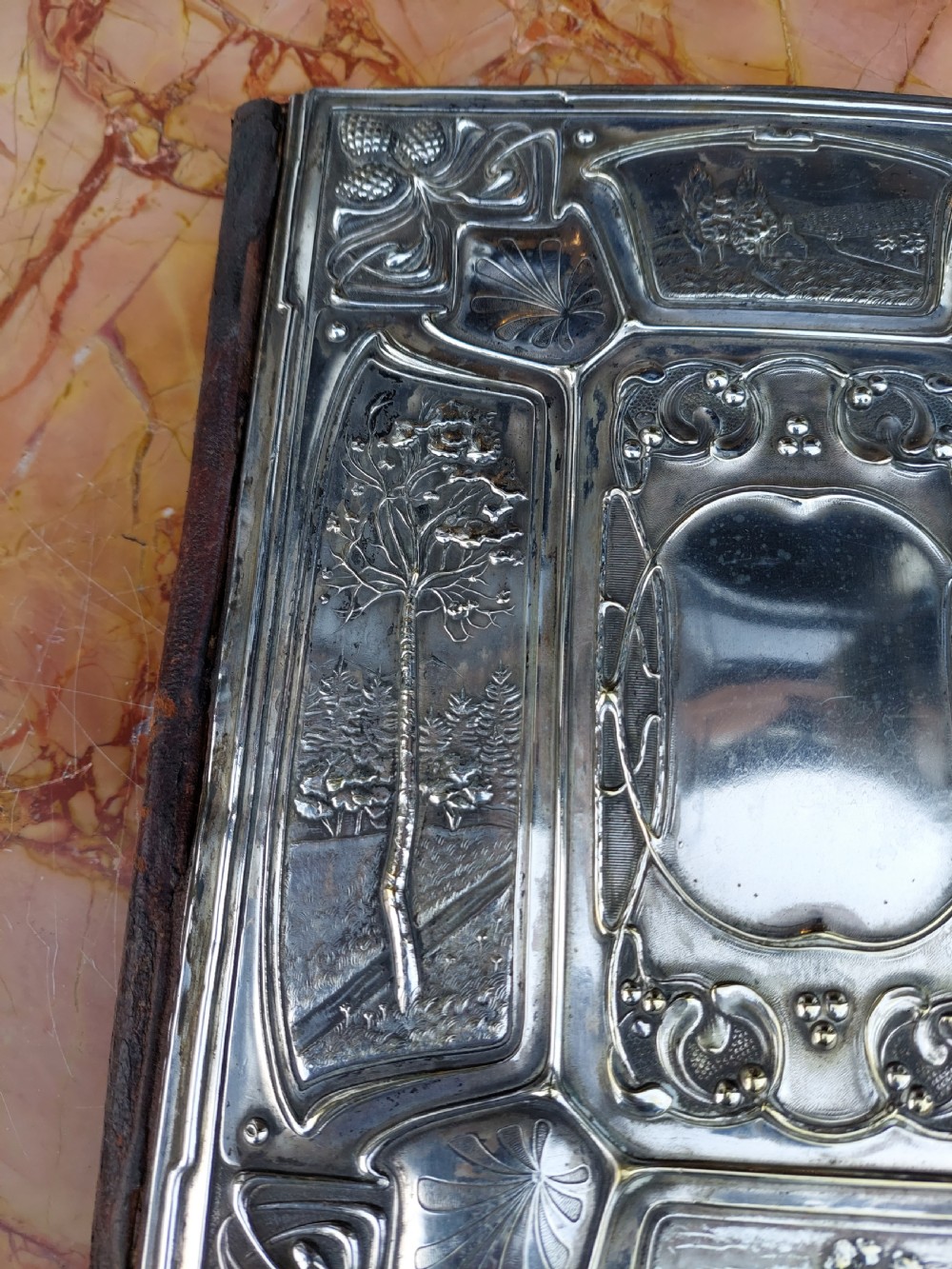 A rectangular silver-plated Art Nouveau blotter folio featuring relief embossed panels of trees, landscapes, pinecone motifs, and a central polished cartouche. View X