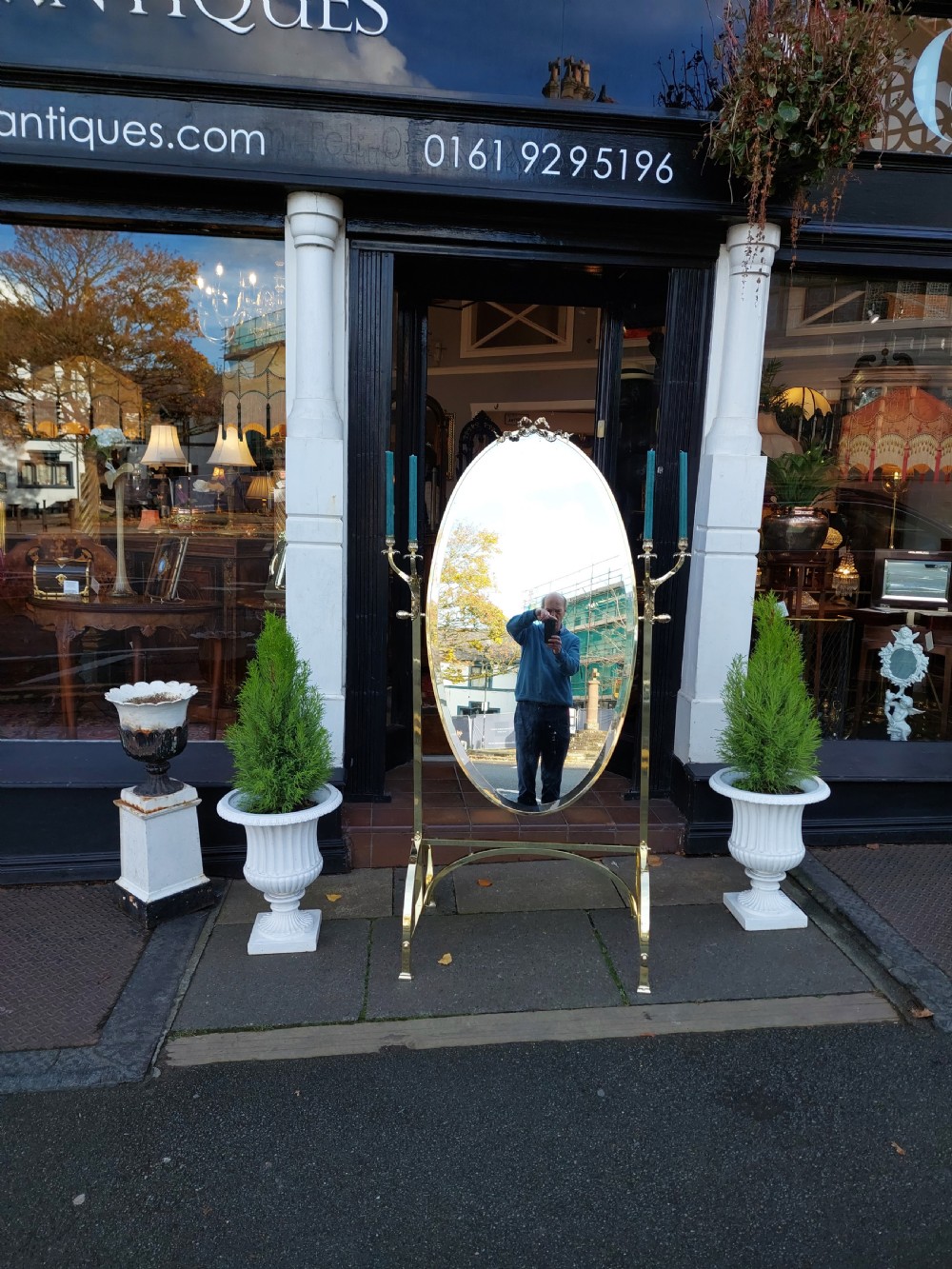 An Edwardian brass cheval mirror featuring an oval bevelled glass frame with decorative cresting and side candle sconces supported on a slender standing base. View IV