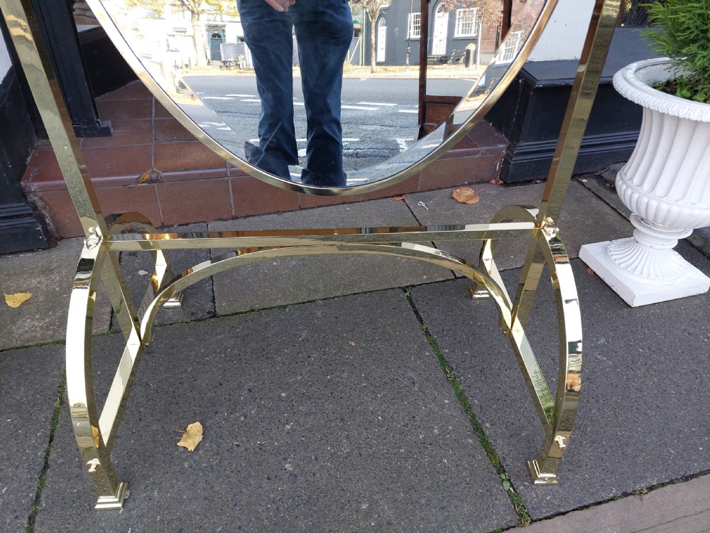 An Edwardian brass cheval mirror featuring an oval bevelled glass frame with decorative cresting and side candle sconces supported on a slender standing base. View V