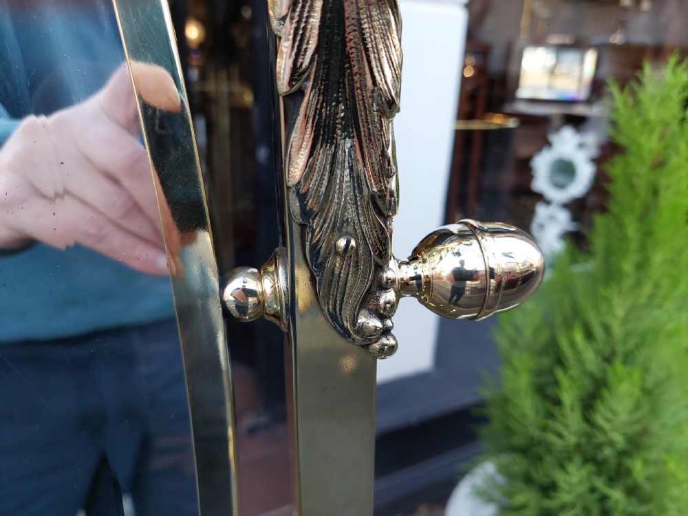 An Edwardian brass cheval mirror featuring an oval bevelled glass frame with decorative cresting and side candle sconces supported on a slender standing base. View IX