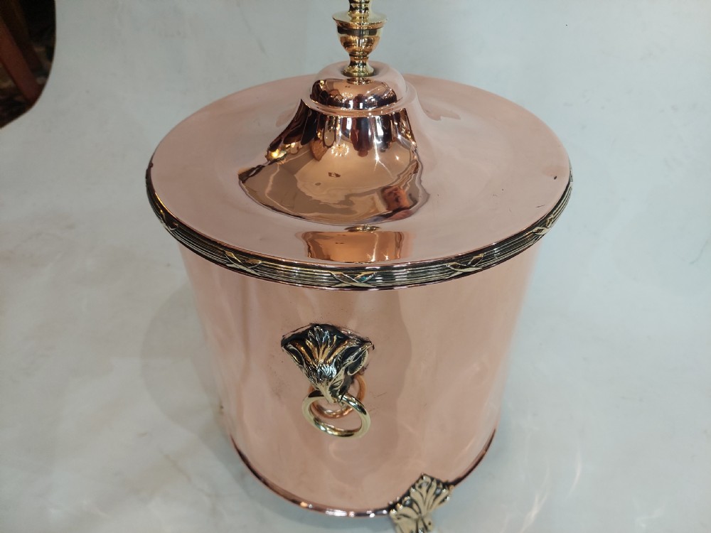 A cylindrical polished copper coal bucket featuring a fitted lid with a brass finial, side ring handles attached to lion mask mounts, and four detailed brass paw feet. View II
