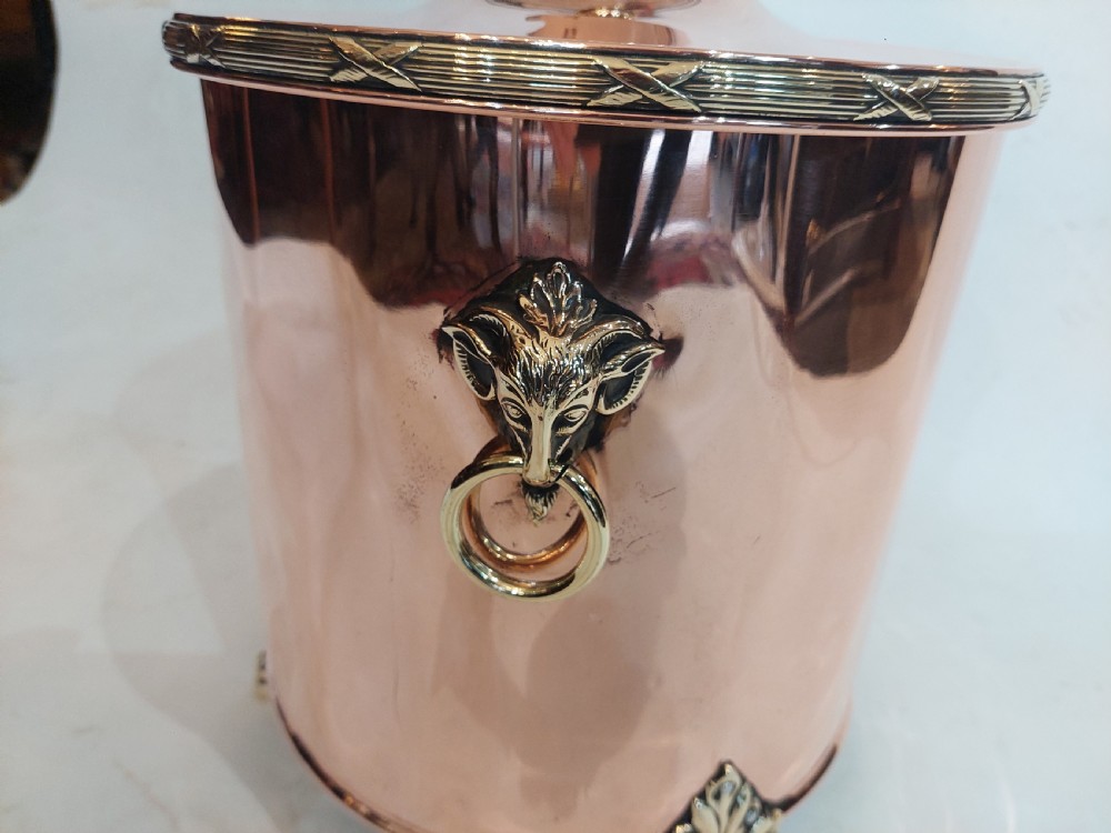 A cylindrical polished copper coal bucket featuring a fitted lid with a brass finial, side ring handles attached to lion mask mounts, and four detailed brass paw feet. View IV