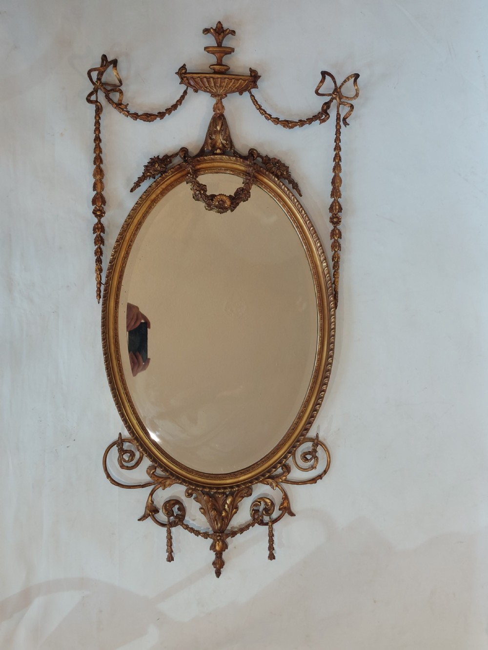 A late Victorian Adam Revival giltwood oval wall mirror decorated with an urn cresting, trailing ribbon swags, and scrollwork accents. View II