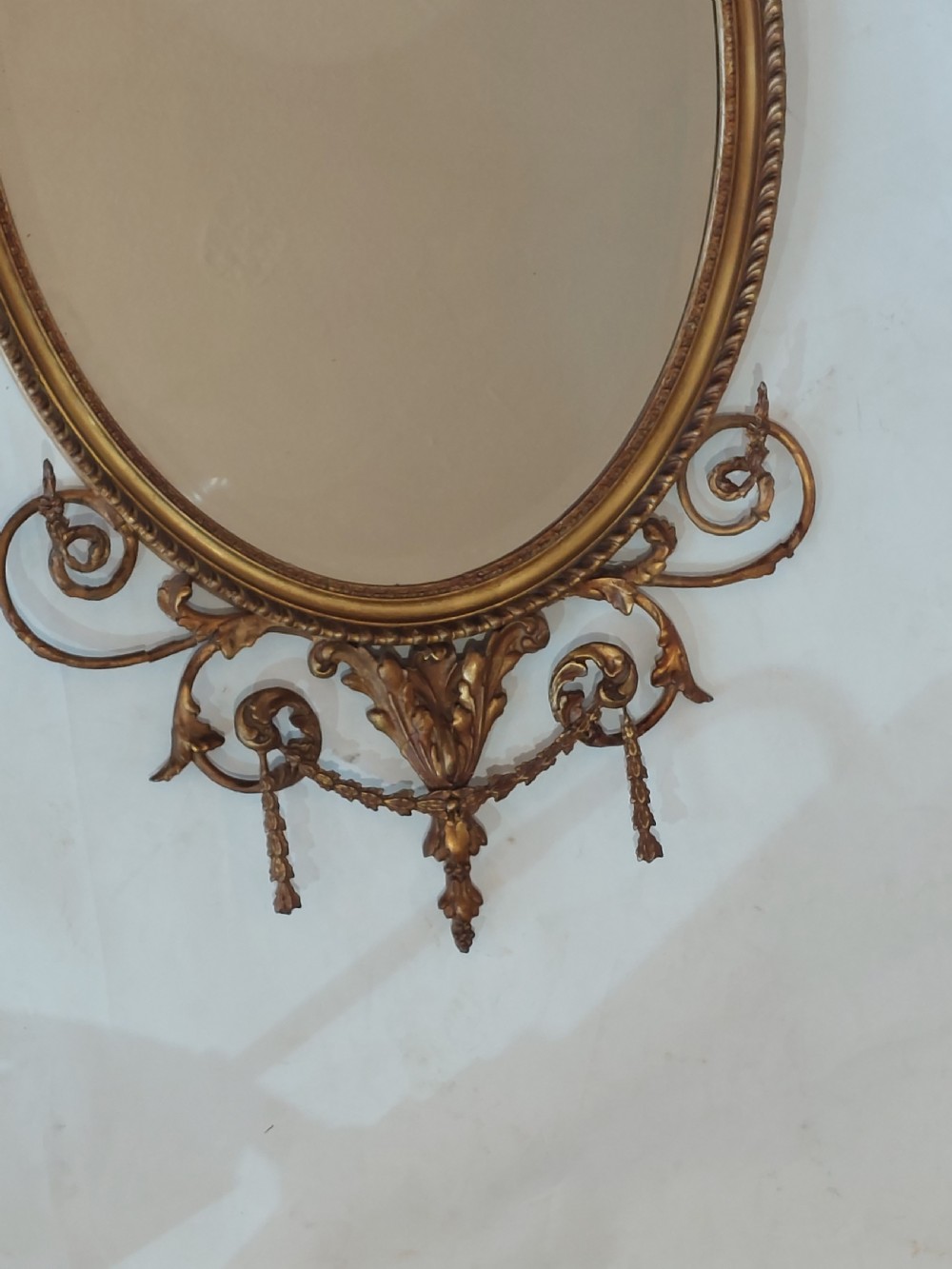 A late Victorian Adam Revival giltwood oval wall mirror decorated with an urn cresting, trailing ribbon swags, and scrollwork accents. View III