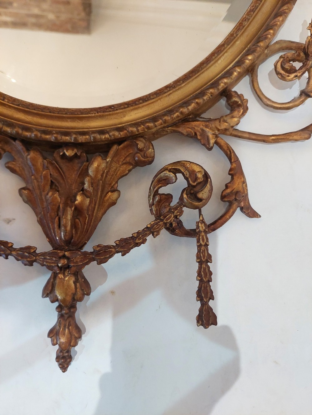 A late Victorian Adam Revival giltwood oval wall mirror decorated with an urn cresting, trailing ribbon swags, and scrollwork accents. View VI