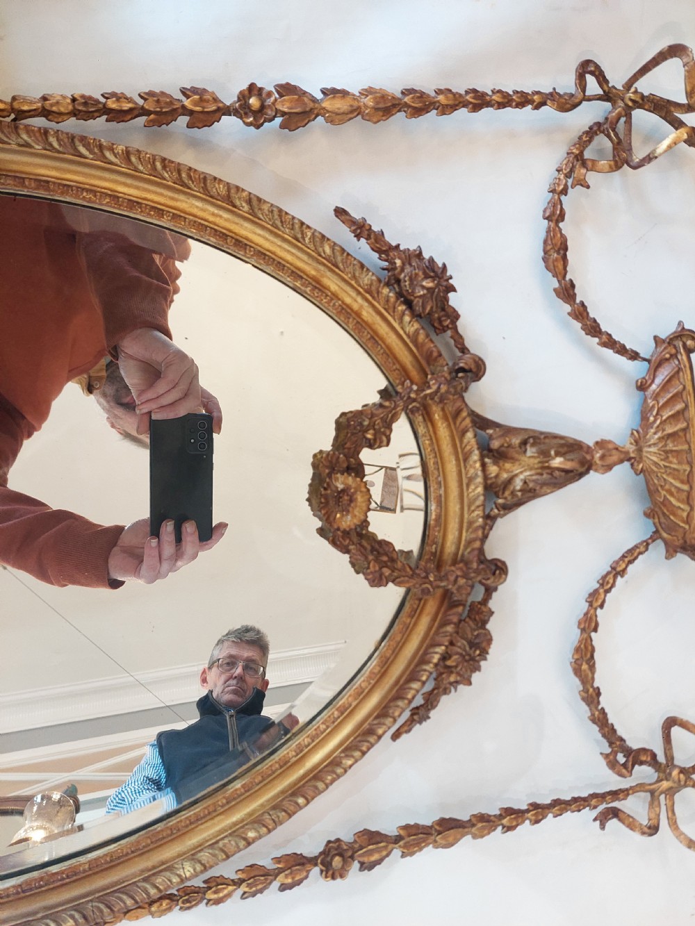 A late Victorian Adam Revival giltwood oval wall mirror decorated with an urn cresting, trailing ribbon swags, and scrollwork accents. View X