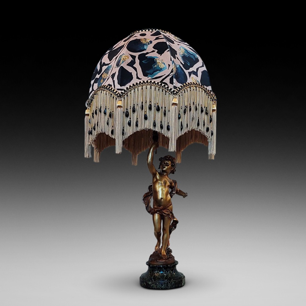 A cast bronze cherub statuette holding aloft a dome-shaped lampshade with patterned fabric and beaded tassel fringe, set on a dark marble base.