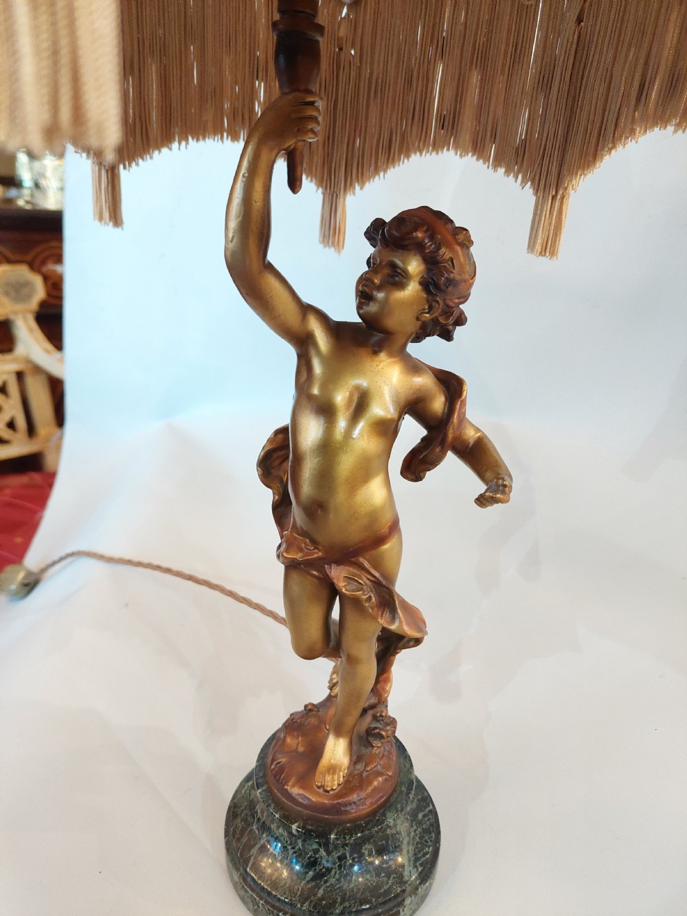 A cast bronze cherub statuette holding aloft a dome-shaped lampshade with patterned fabric and beaded tassel fringe, set on a dark marble base. View IV
