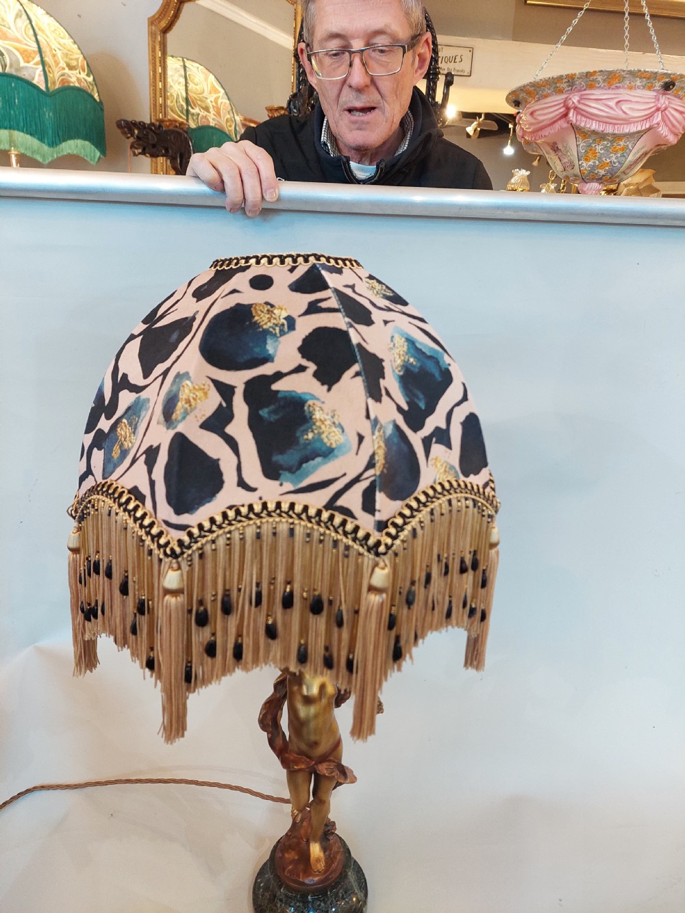 A cast bronze cherub statuette holding aloft a dome-shaped lampshade with patterned fabric and beaded tassel fringe, set on a dark marble base. View VI