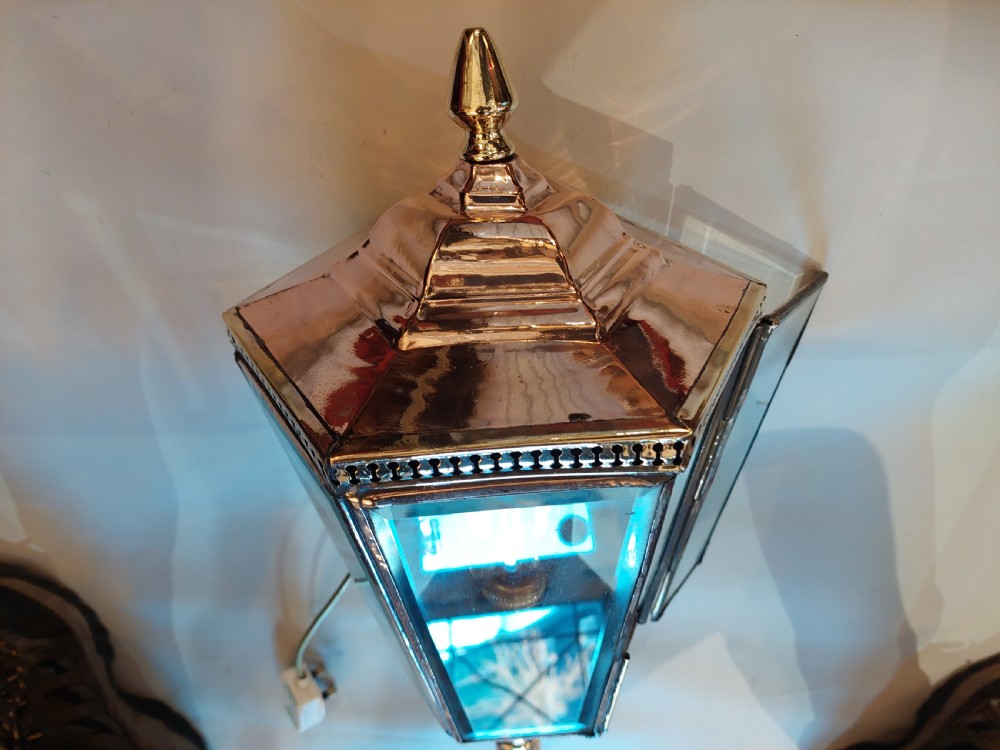 An Edwardian hexagonal wall-mounted porch lantern featuring polished brass and copper framing, clear glass panels, a pointed top finial, and a turned lower stem. View VII