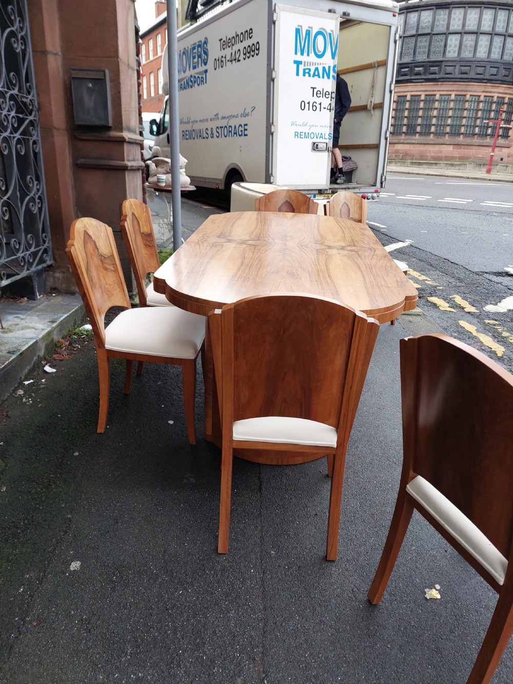 An Art Deco walnut dining suite comprising an oval pedestal table and six matching chairs with arched burr walnut backs and cream upholstered seats. View III
