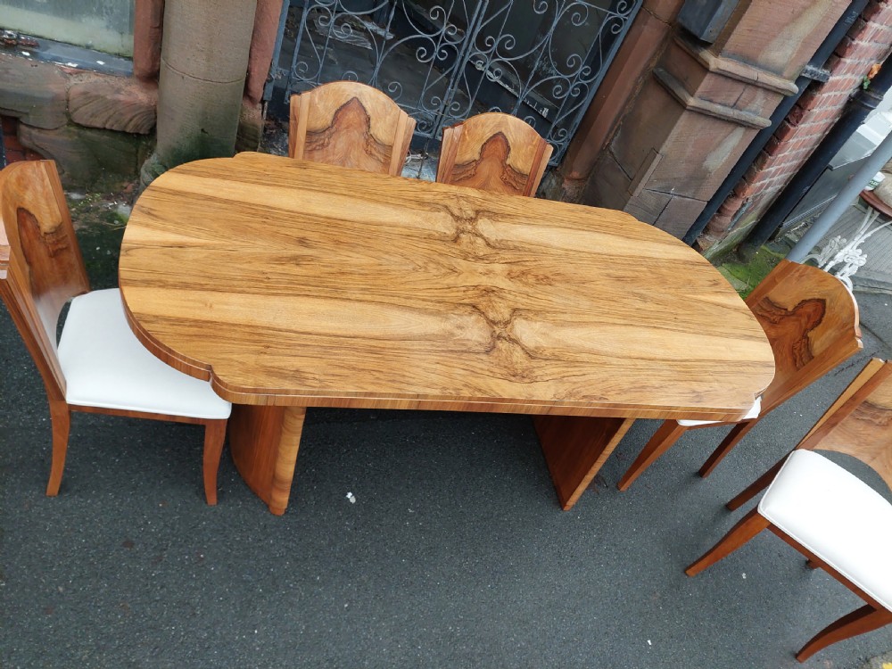 An Art Deco walnut dining suite comprising an oval pedestal table and six matching chairs with arched burr walnut backs and cream upholstered seats. View VI