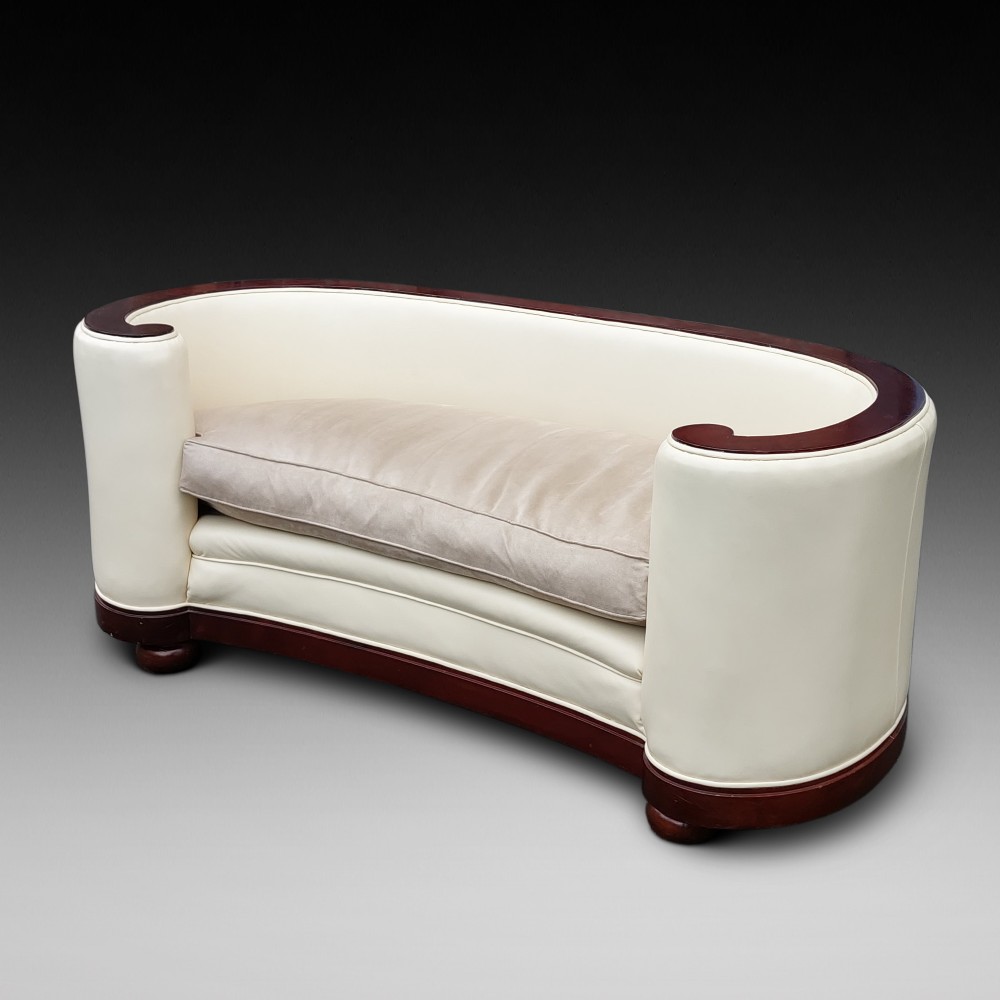 A curved Art Deco sofa featuring cream leather upholstery, a soft seat cushion, and polished palisander wood scroll trims on a low wooden base.