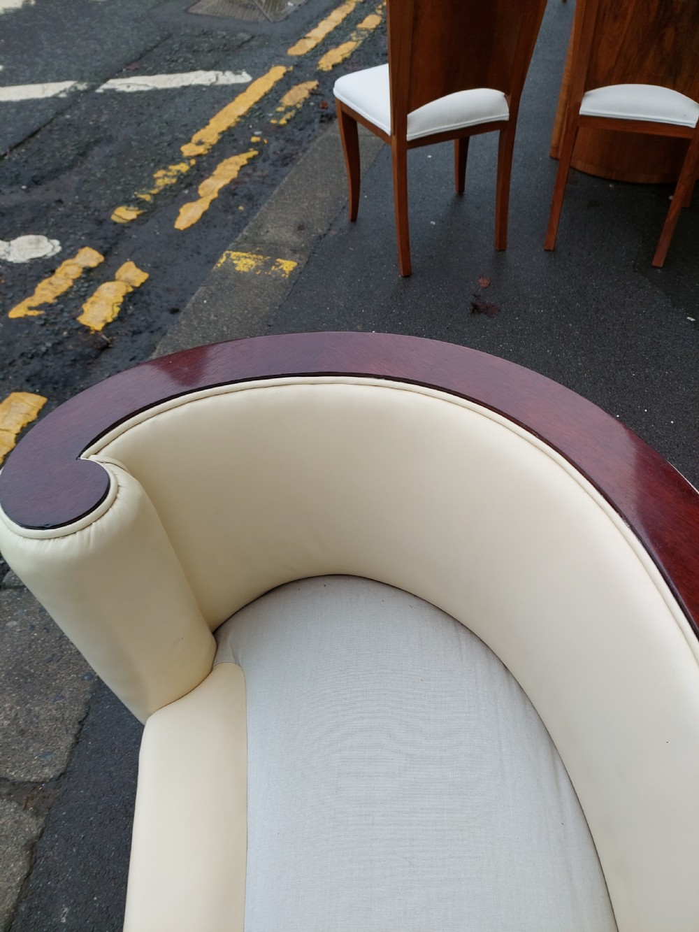 A curved Art Deco sofa featuring cream leather upholstery, a soft seat cushion, and polished palisander wood scroll trims on a low wooden base. View V