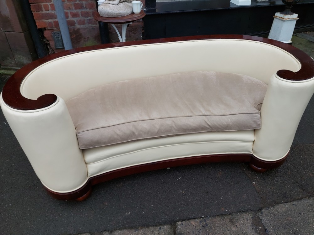 A curved Art Deco sofa featuring cream leather upholstery, a soft seat cushion, and polished palisander wood scroll trims on a low wooden base. View VI