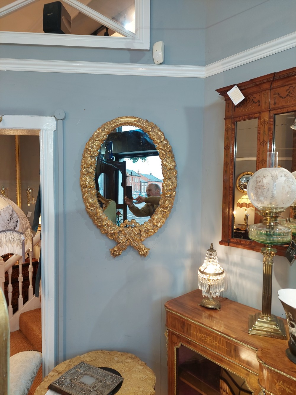 An oval Victorian wall mirror featuring a giltwood frame carved with floral motifs and a tied ribbon detail at the base. View II