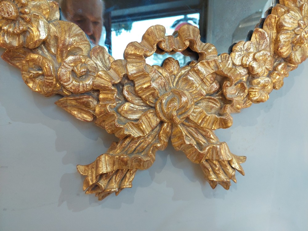 An oval Victorian wall mirror featuring a giltwood frame carved with floral motifs and a tied ribbon detail at the base. View III