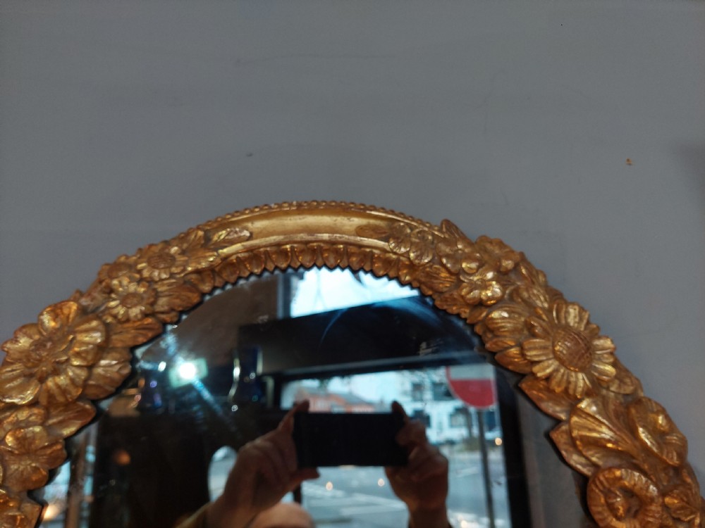 An oval Victorian wall mirror featuring a giltwood frame carved with floral motifs and a tied ribbon detail at the base. View V