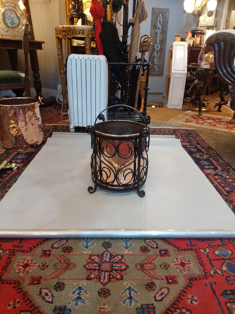 A cylindrical copper bucket set within a black wrought iron frame featuring scroll motifs and a curved carrying handle. View II