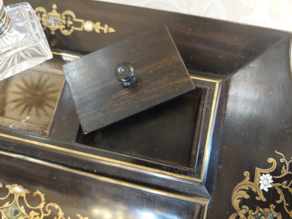 A mid-19th-century dark coromandel wood inkstand desk tidy featuring floral inlay, a pen trough, two lidded compartments, and a central glass inkwell. View VI