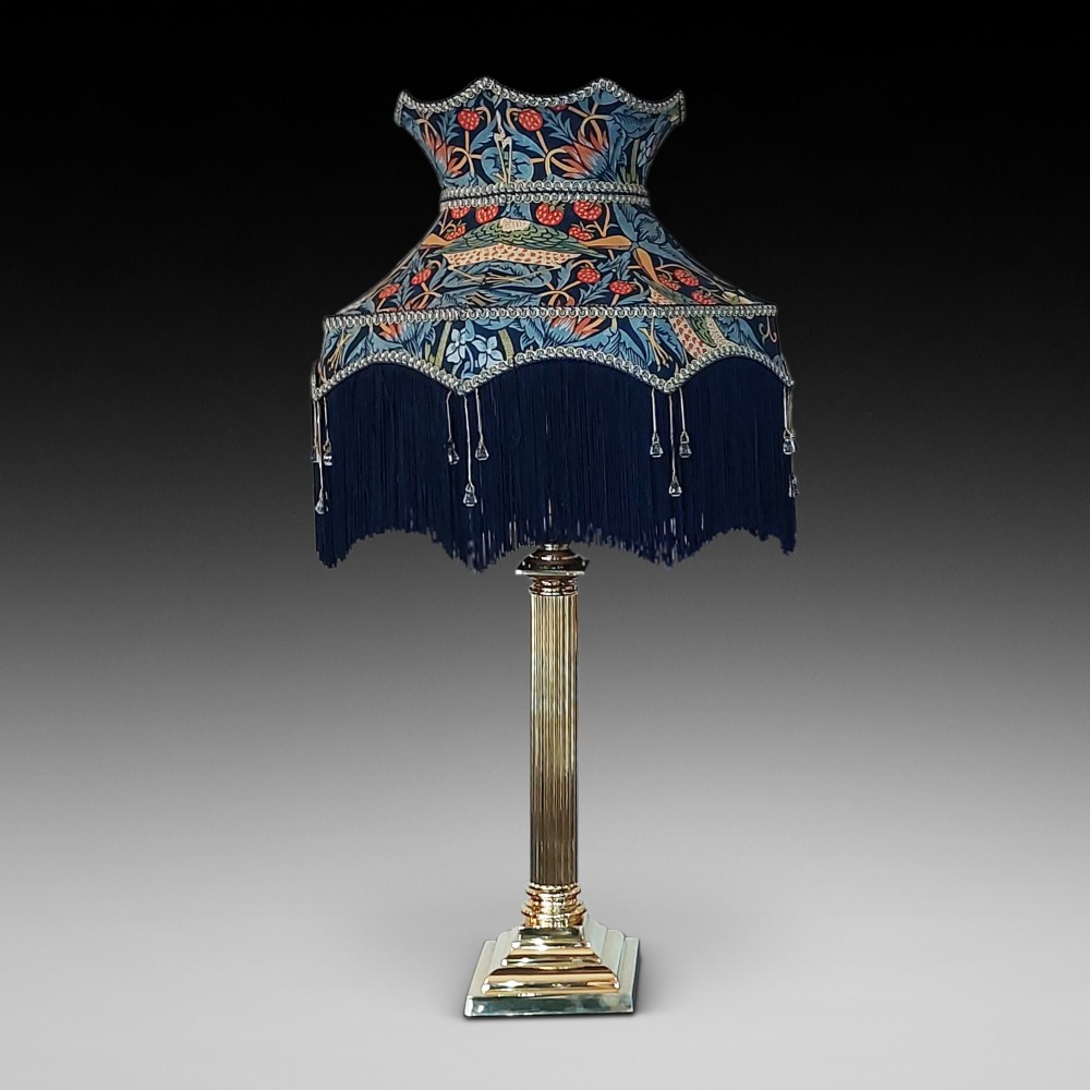 An early 20th-century brass table lamp featuring a reeded Doric column base and a patterned fabric lampshade with dark blue fringe and crystal drop beads.