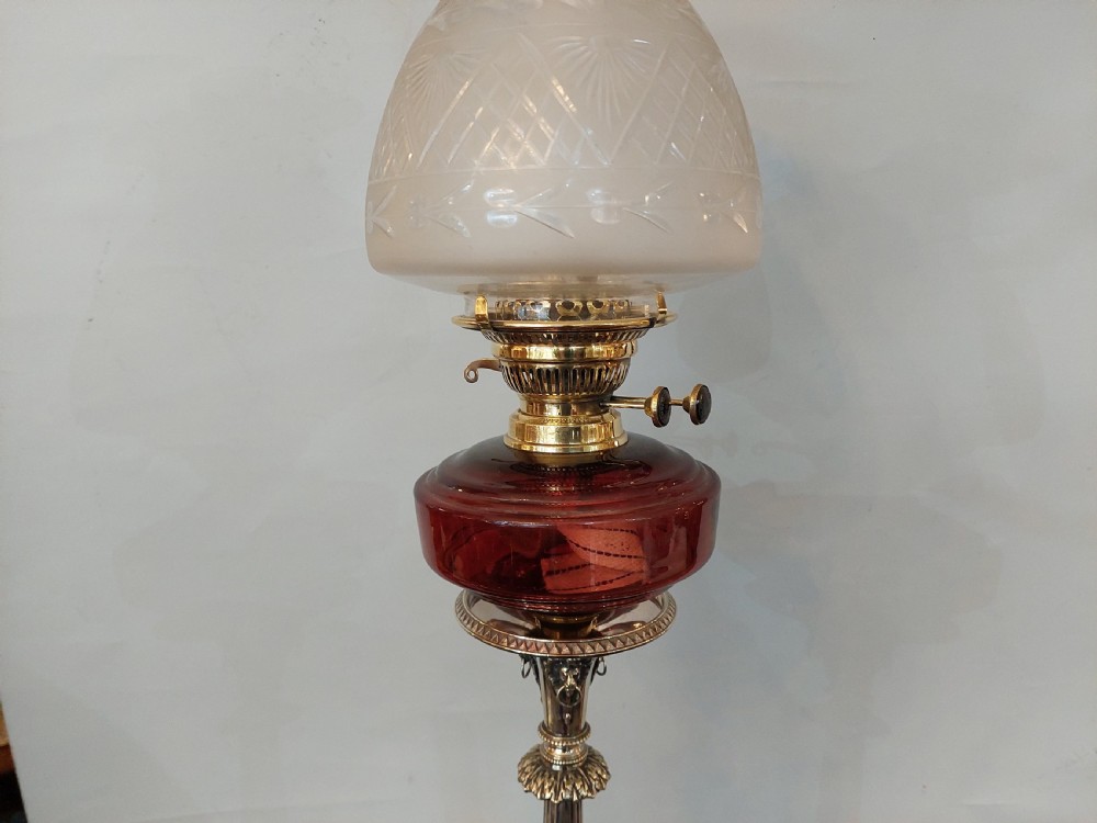 A late Victorian silver-plated oil lamp featuring a fluted column stem, a red cranberry glass reservoir, a brass burner, and a frosted etched glass shade. View VI