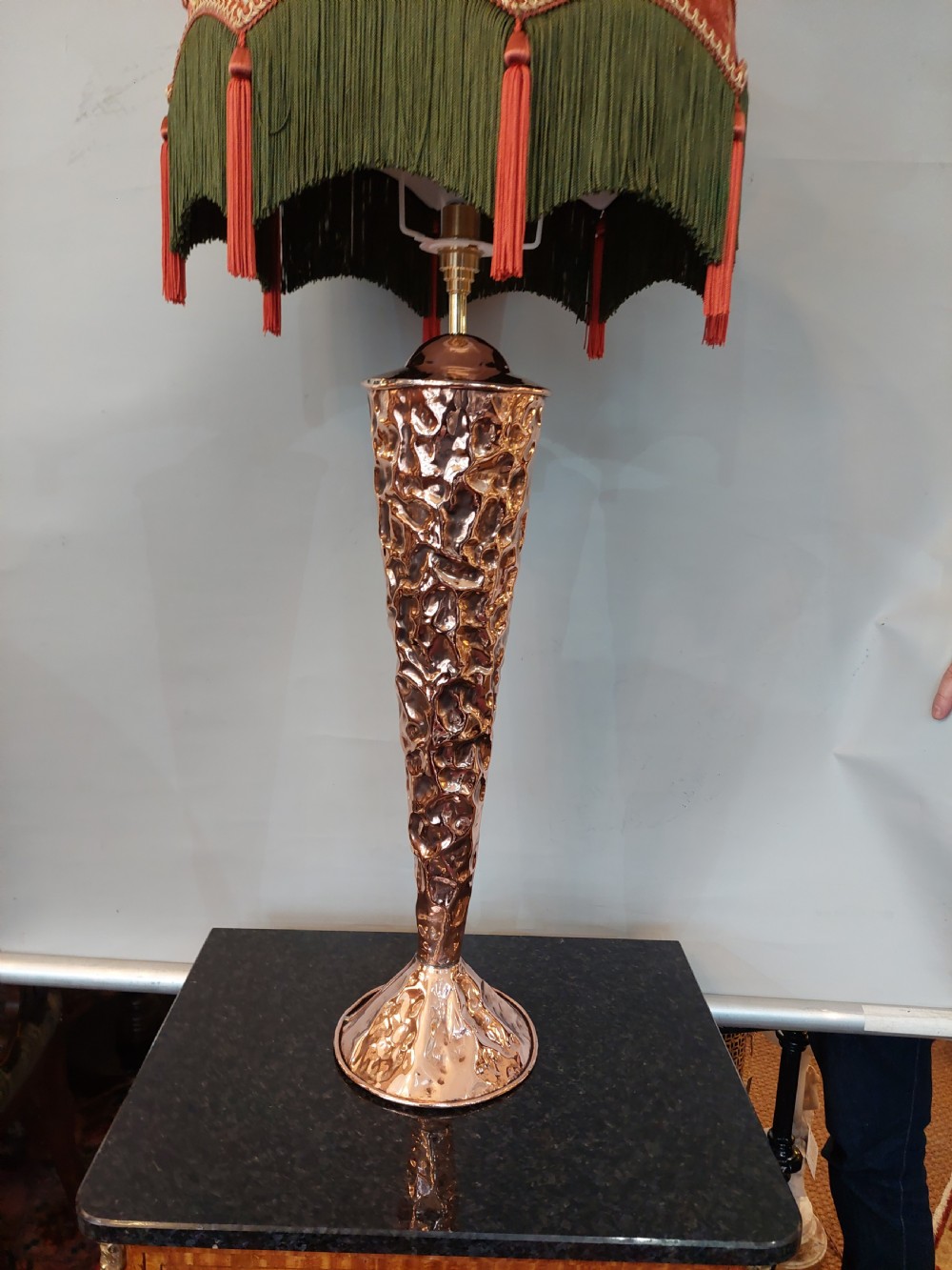 An Arts and Crafts style table lamp featuring a tall, tapered beaten copper base topped with a decorative fringed fabric lampshade. View III