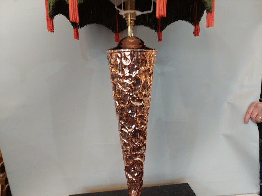 An Arts and Crafts style table lamp featuring a tall, tapered beaten copper base topped with a decorative fringed fabric lampshade. View V