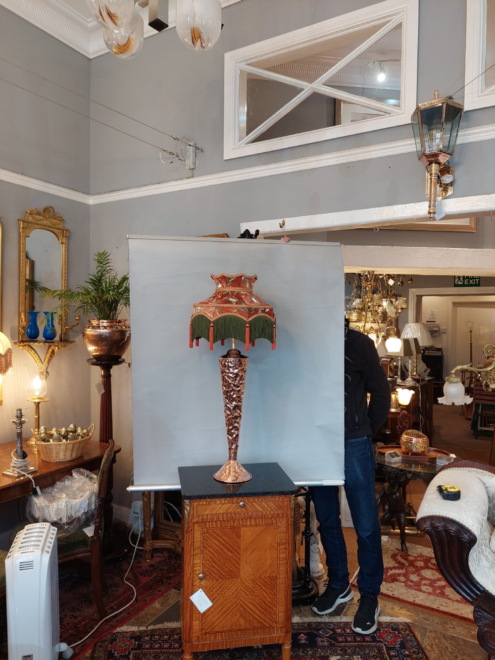 An Arts and Crafts style table lamp featuring a tall, tapered beaten copper base topped with a decorative fringed fabric lampshade. View VI