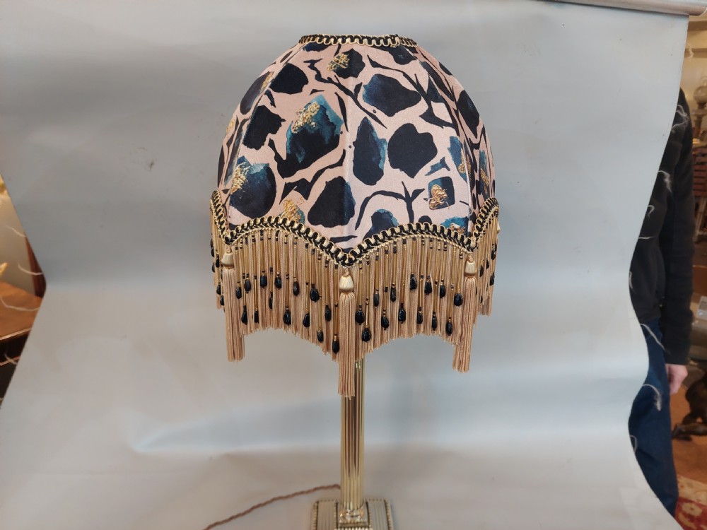 An early twentieth-century brass table lamp featuring a fluted Corinthian column shaft on a stepped pedestal base, topped with a patterned fabric shade with tassel and bead fringe. View IV