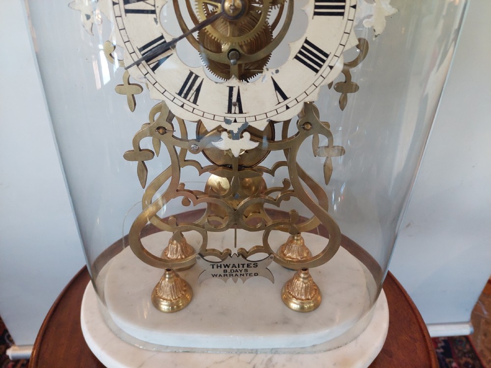 A Victorian brass skeleton clock with openwork gear movement, silvered chapter ring, and top bell, enclosed under a clear glass dome on a white marble base. View IV