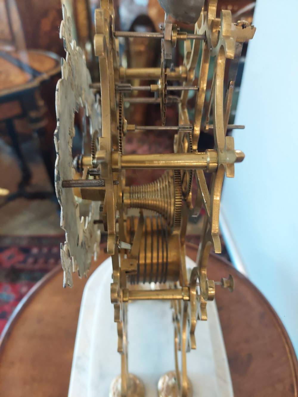 A Victorian brass skeleton clock with openwork gear movement, silvered chapter ring, and top bell, enclosed under a clear glass dome on a white marble base. View VII