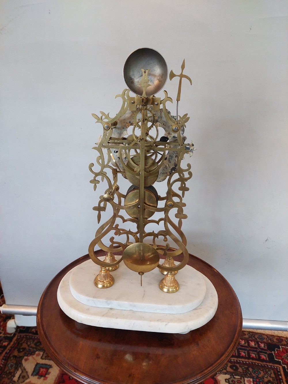 A Victorian brass skeleton clock with openwork gear movement, silvered chapter ring, and top bell, enclosed under a clear glass dome on a white marble base. View VIII