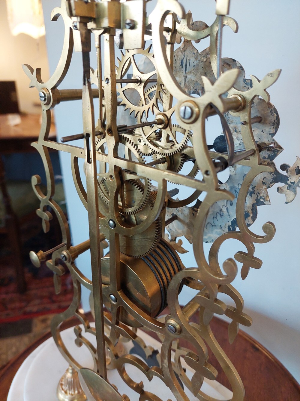A Victorian brass skeleton clock with openwork gear movement, silvered chapter ring, and top bell, enclosed under a clear glass dome on a white marble base. View IX