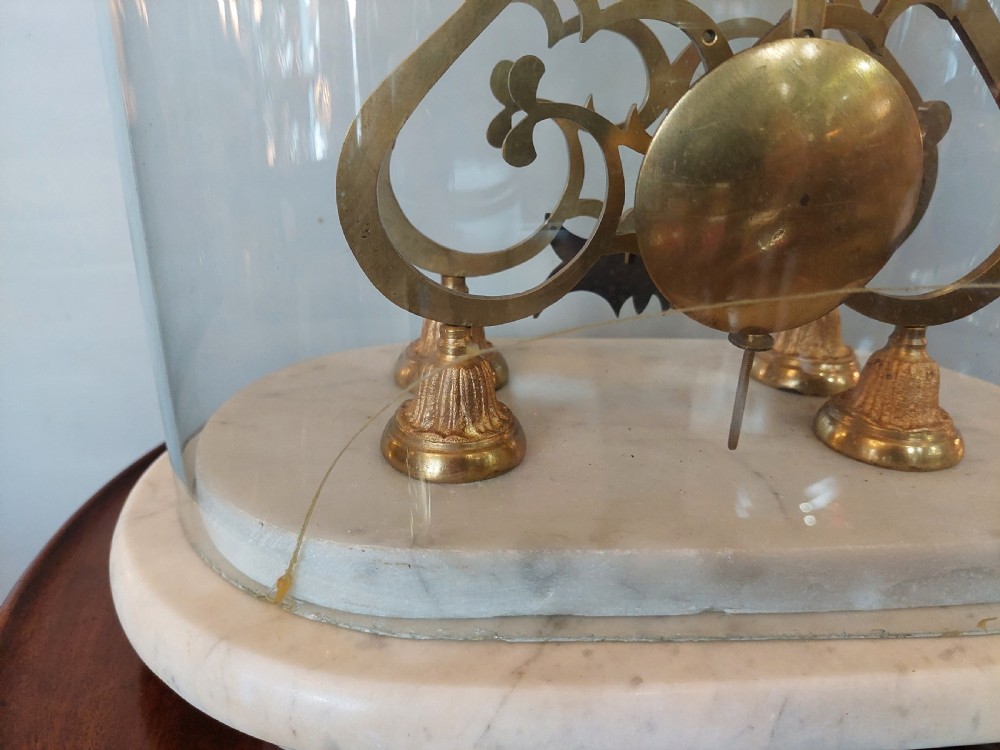 A Victorian brass skeleton clock with openwork gear movement, silvered chapter ring, and top bell, enclosed under a clear glass dome on a white marble base. View X