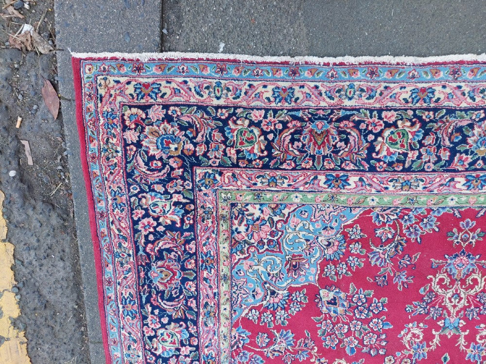 A hand-woven rectangular Persian wool carpet featuring a crimson field with a central floral medallion, pale blue corner spandrels, and a navy blue multi-stripe border. View II
