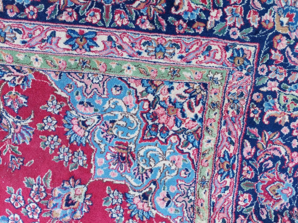 A hand-woven rectangular Persian wool carpet featuring a crimson field with a central floral medallion, pale blue corner spandrels, and a navy blue multi-stripe border. View III