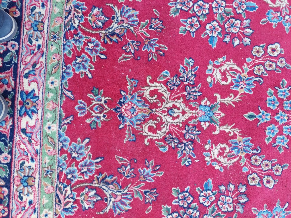 A hand-woven rectangular Persian wool carpet featuring a crimson field with a central floral medallion, pale blue corner spandrels, and a navy blue multi-stripe border. View IV