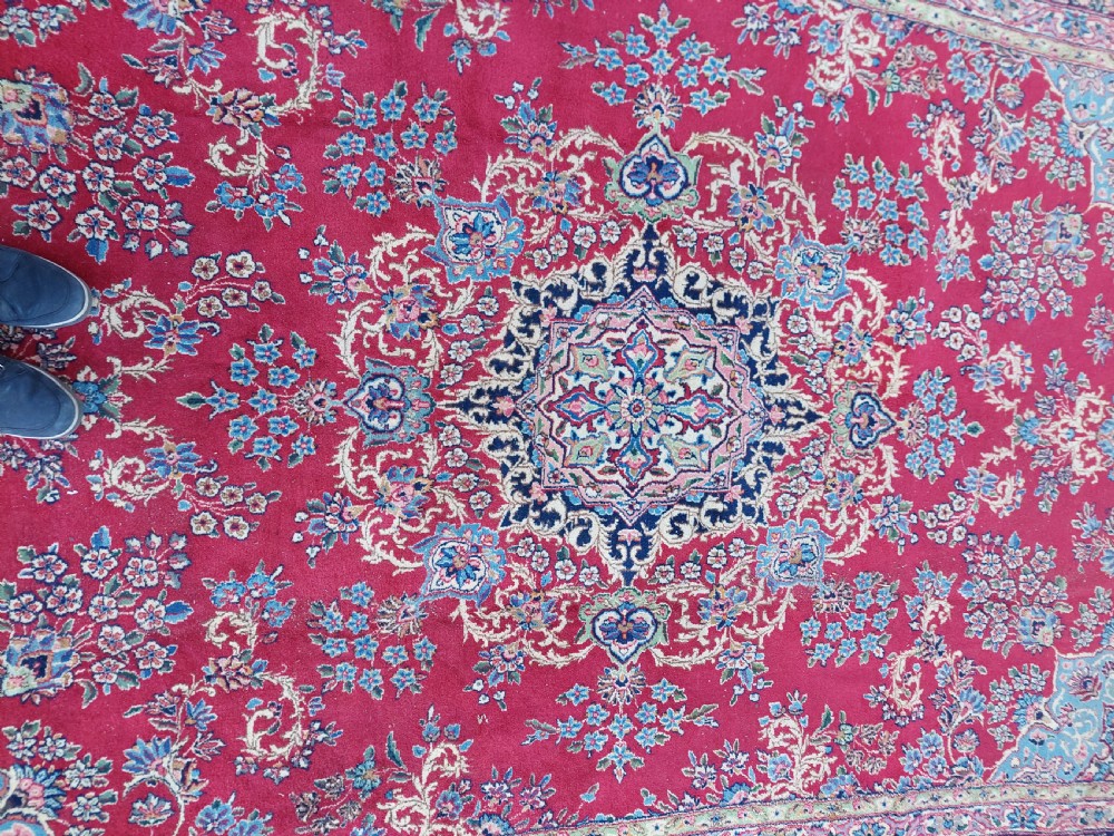 A hand-woven rectangular Persian wool carpet featuring a crimson field with a central floral medallion, pale blue corner spandrels, and a navy blue multi-stripe border. View V