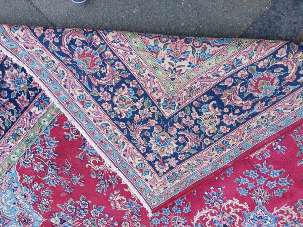 A hand-woven rectangular Persian wool carpet featuring a crimson field with a central floral medallion, pale blue corner spandrels, and a navy blue multi-stripe border. View VI