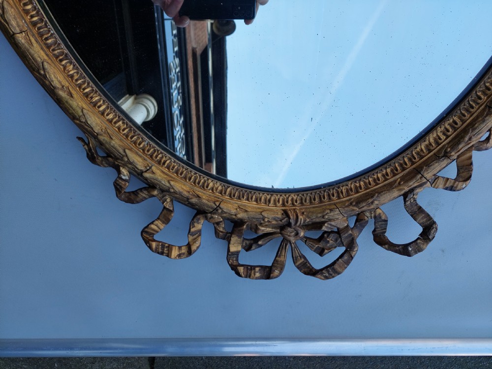 A mid 19th-century oval wall mirror featuring a giltwood frame topped with a carved ribbon bow cresting. View IV