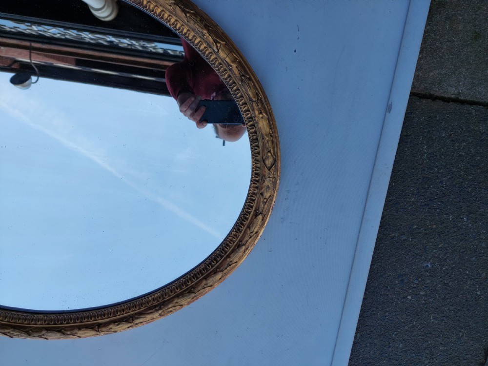 A mid 19th-century oval wall mirror featuring a giltwood frame topped with a carved ribbon bow cresting. View V
