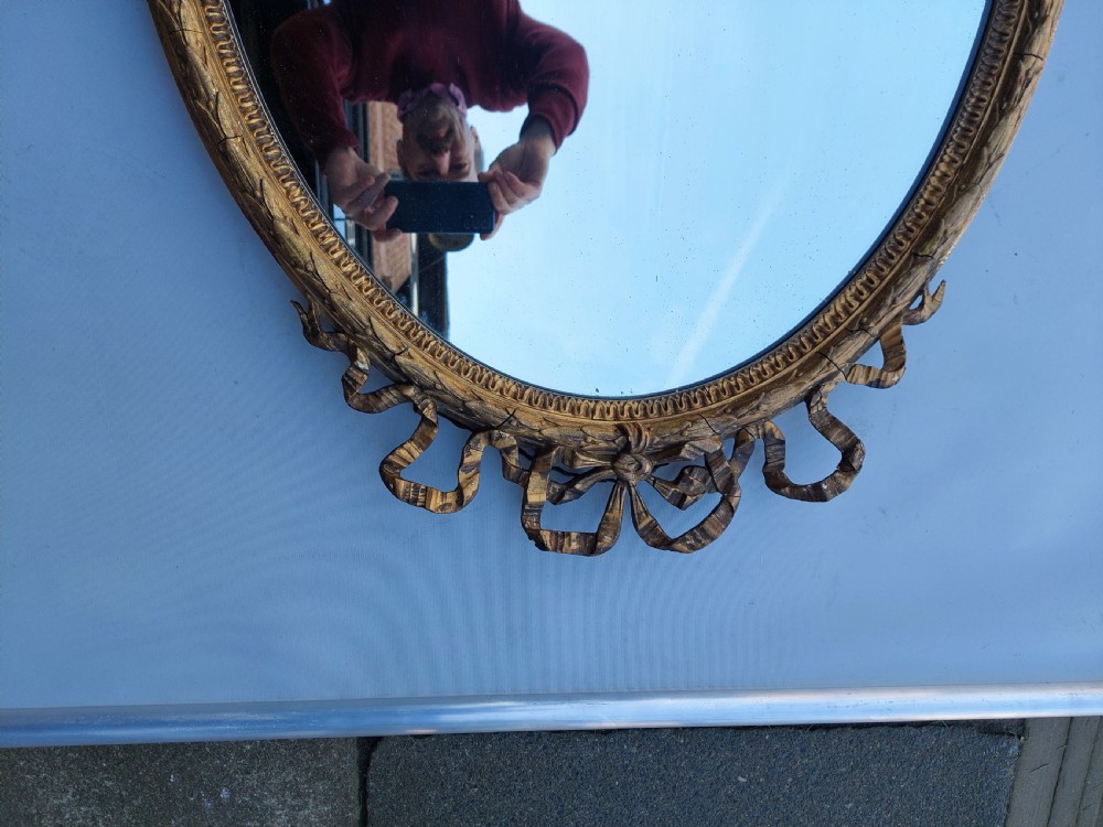 A mid 19th-century oval wall mirror featuring a giltwood frame topped with a carved ribbon bow cresting. View VI