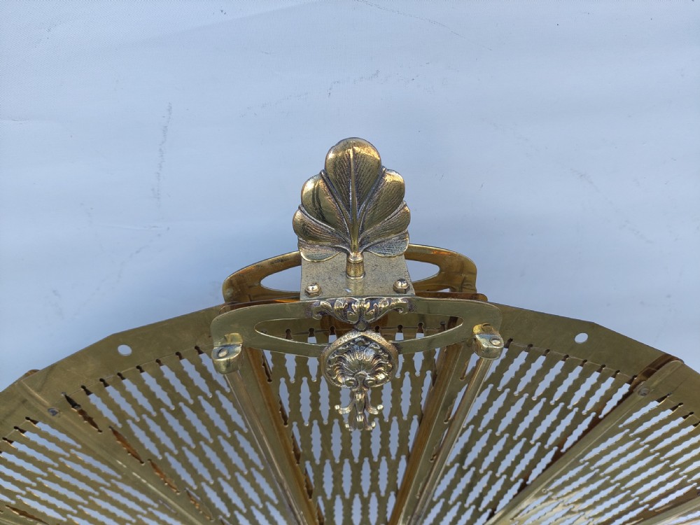 A Victorian folding brass fireplace screen shaped like an open fan with pierced filigree blades mounted on a cast base. View II