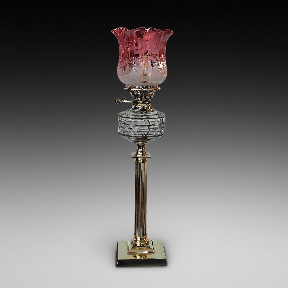 A late 19th-century brass table oil lamp featuring a reeded column, square base, clear glass reservoir, and an etched pink glass shade with a ruffled rim.
